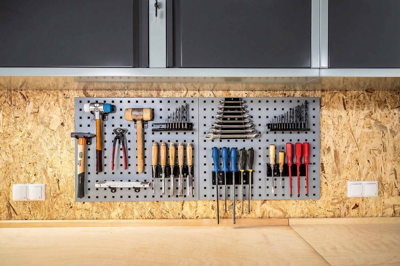 Organisierte Werkzeugwand mit grauen Lochplatten an einer OSB-Wand über einer Werkbank, mit Hämmern, Zangen, Beiteln und Schraubendrehern.
