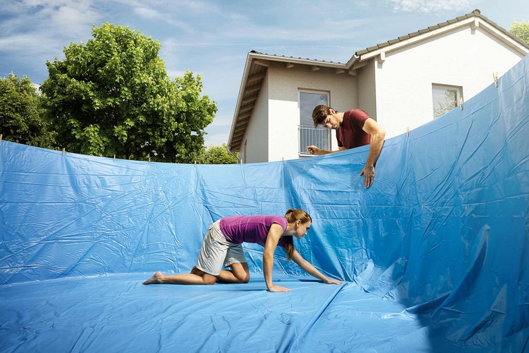 Scène avec deux personnes installant une bâche de piscine bleue, avec une maison et des arbres en arrière-plan.