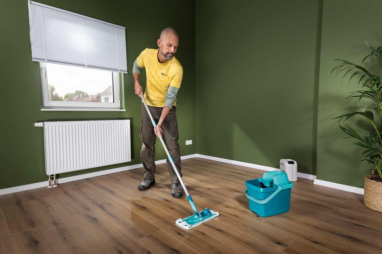 Un homme lave un plancher en bois avec une serpillière dans une pièce avec une fenêtre, un radiateur et une plante en pot