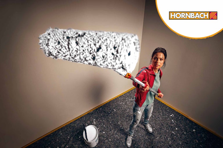 Frau streicht mit einer Farbwalze eine Wand, Eimer mit Farbe steht im Raum, Hornbach Logo