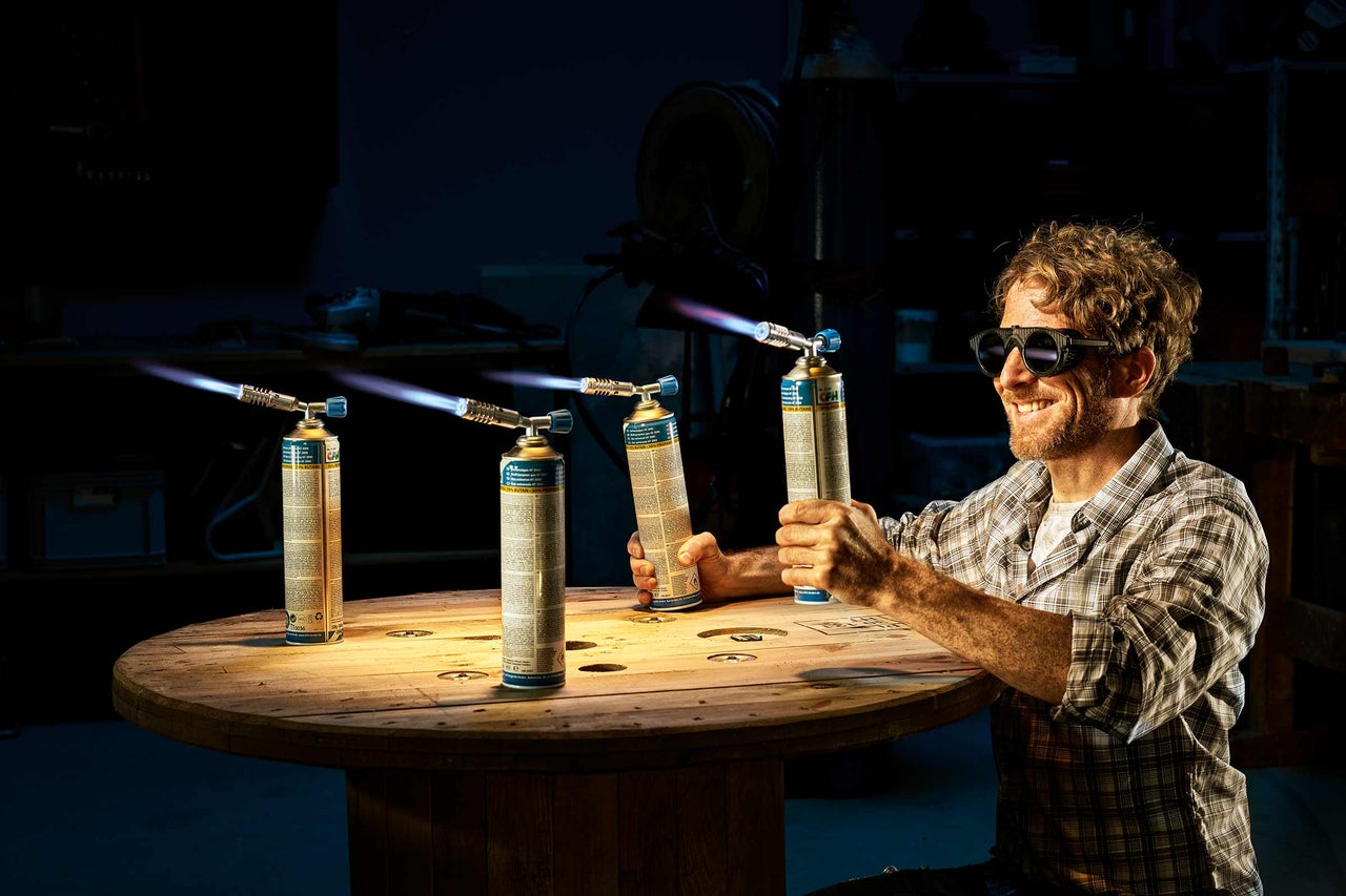 Un homme portant des lunettes de protection tient un brûleur à gaz dans sa main, tandis que trois autres brûleurs à gaz sont posés allumés sur une table en bois.