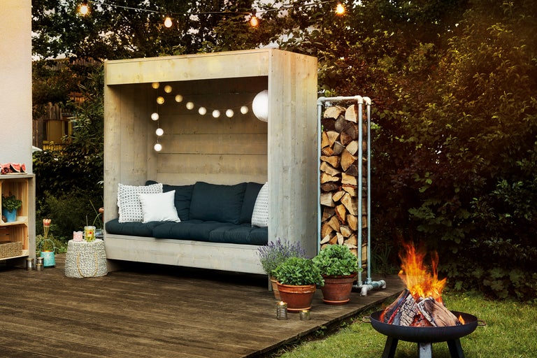 Ambiance de jardin conviviale avec terrasse en bois, coin salon avec coussins, pile de bois, pots de fleurs et foyer