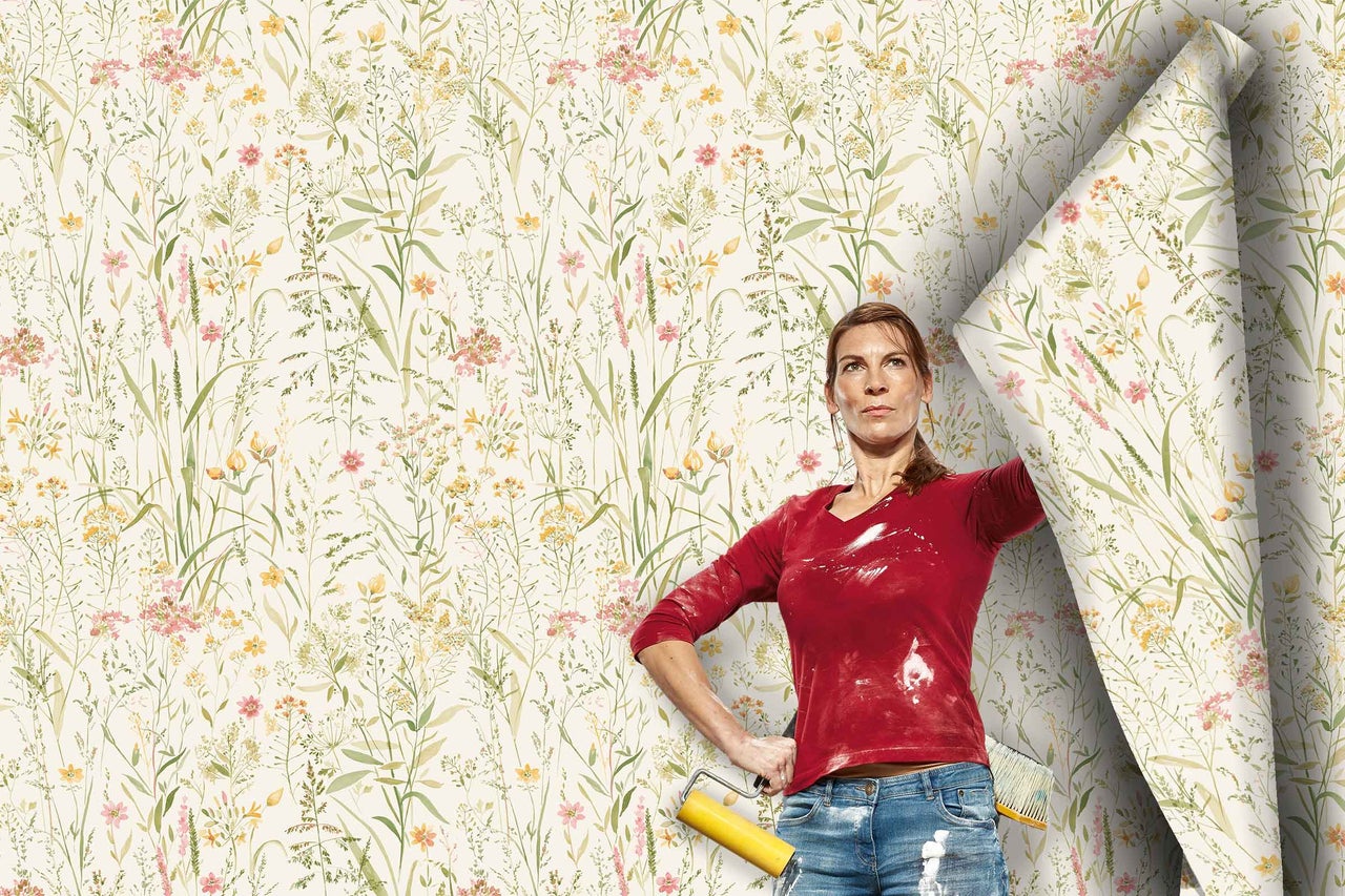 Une femme avec des outils de tapisserie se tient devant un mur en cours de tapissage avec un papier peint intissé à motif de prairie fleurie.