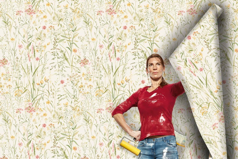 Une femme avec des outils de tapisserie se tient devant un mur en cours de tapissage avec un papier peint intissé à motif de prairie fleurie.