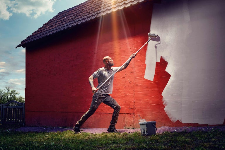 Un homme peint un mur de maison rouge en blanc avec un rouleau à peinture