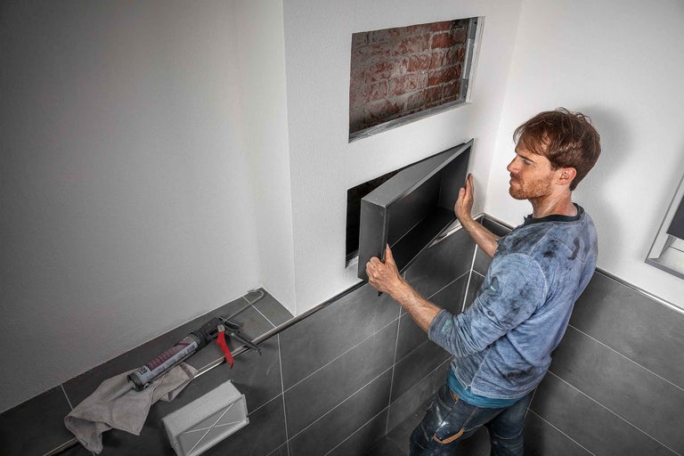 Handwerker montiert eine Revisionsklappe in einer Badezimmerwand. Werkzeuge liegen bereit.