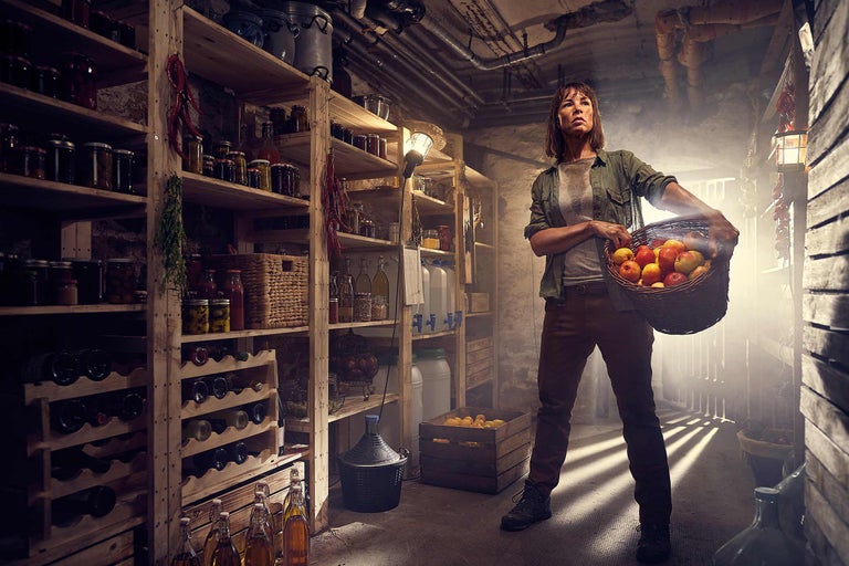 Une femme se tient dans une cave avec des étagères remplies de conserves, de bouteilles et de provisions et tient un panier de pommes.
