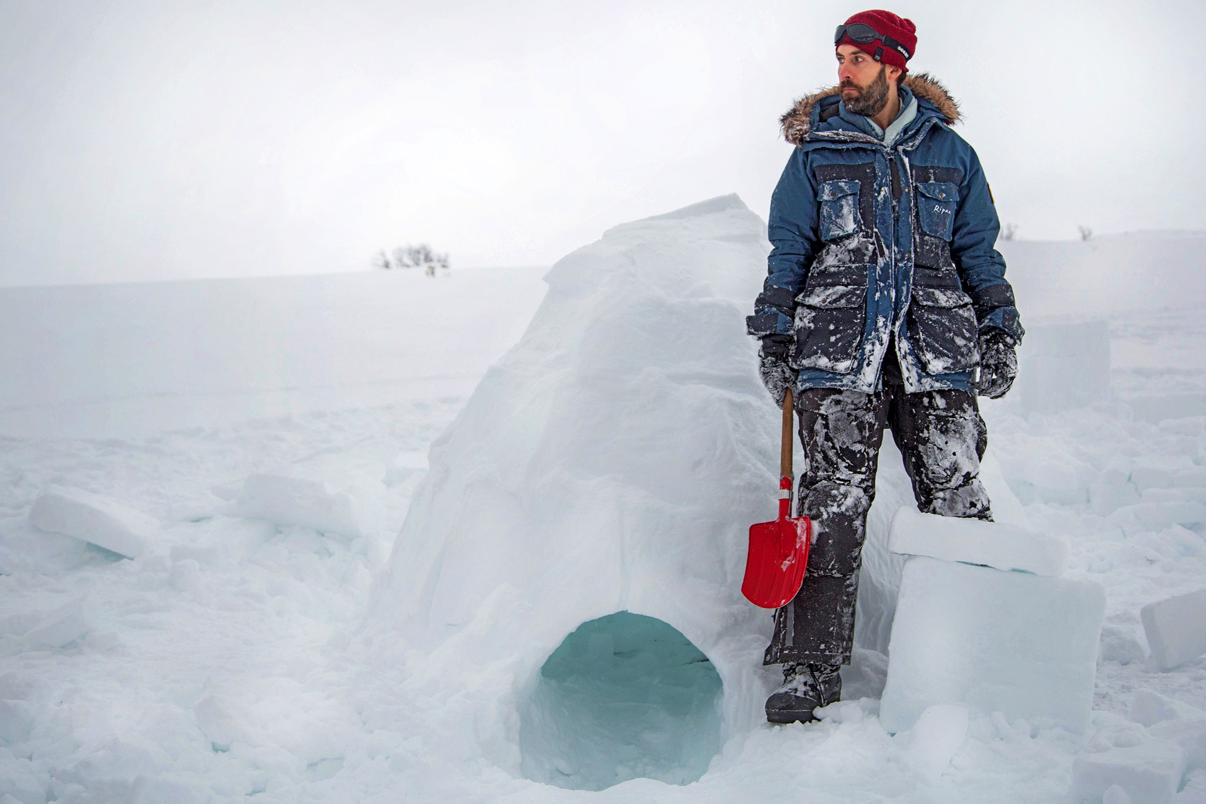 Mann in Winterkleidung steht neben einem Iglu mit einer Schaufel im Schnee.