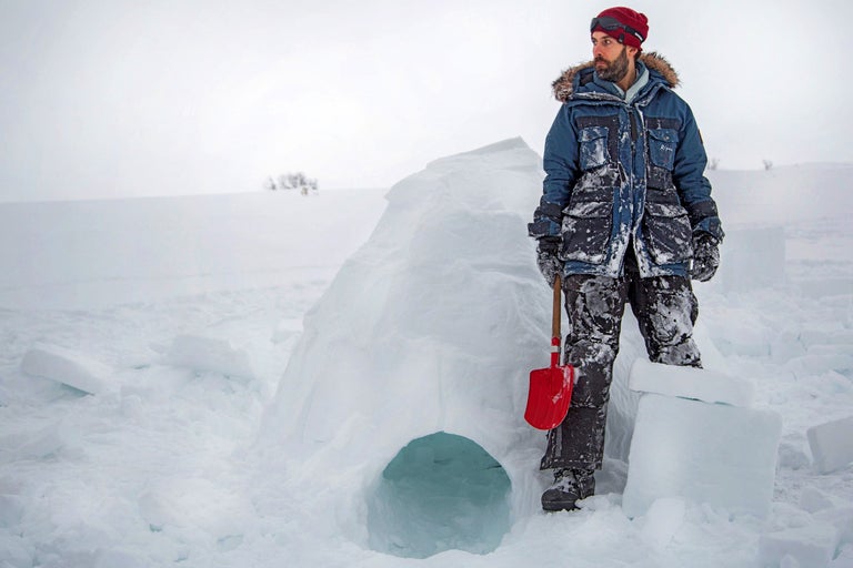 Homme en vêtements d'hiver debout à côté d'un igloo avec une pelle dans la neige.