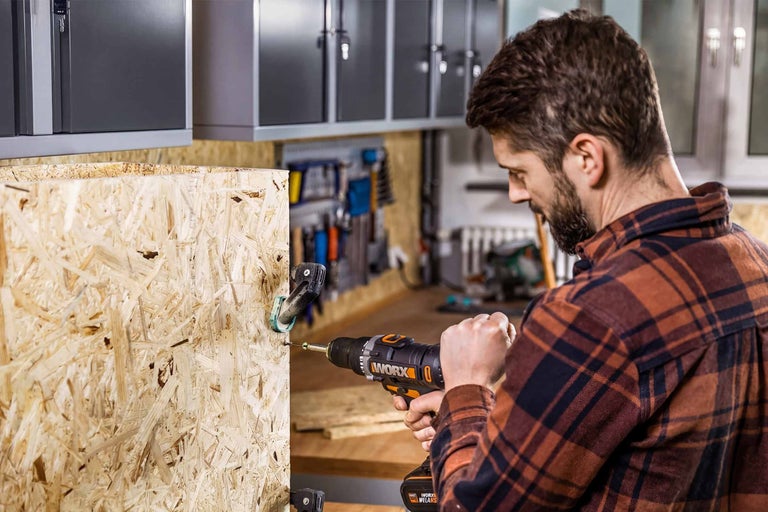 Mann nutzt Worx Akku-Bohrschrauber an OSB-Platte in Werkstatt mit Wandschränken und Werkzeughaltern.