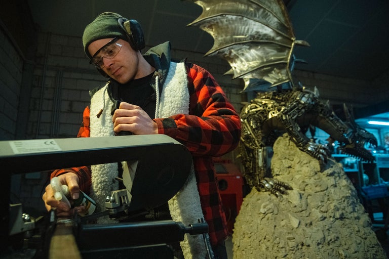 Mann mit Schutzbrille und Gehörschutz, der eine Maschine bedient, im Hintergrund eine Metalldrache Skulptur.