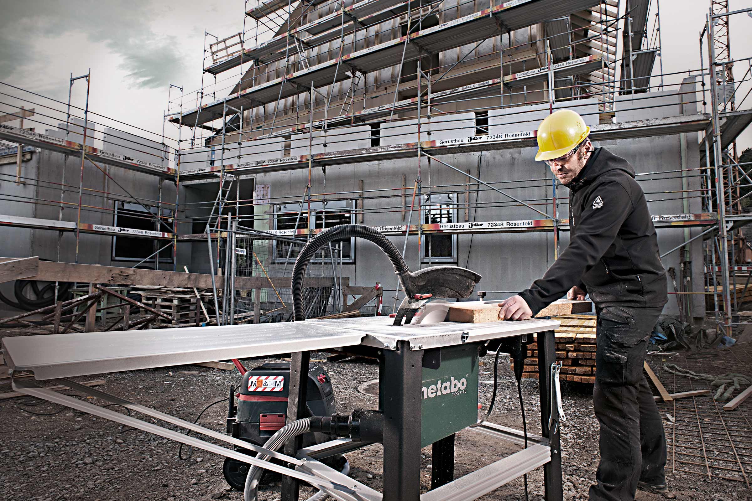 Un ouvrier du bâtiment avec un casque de protection coupe une planche de bois avec une scie circulaire sur table Metabo sur un chantier.