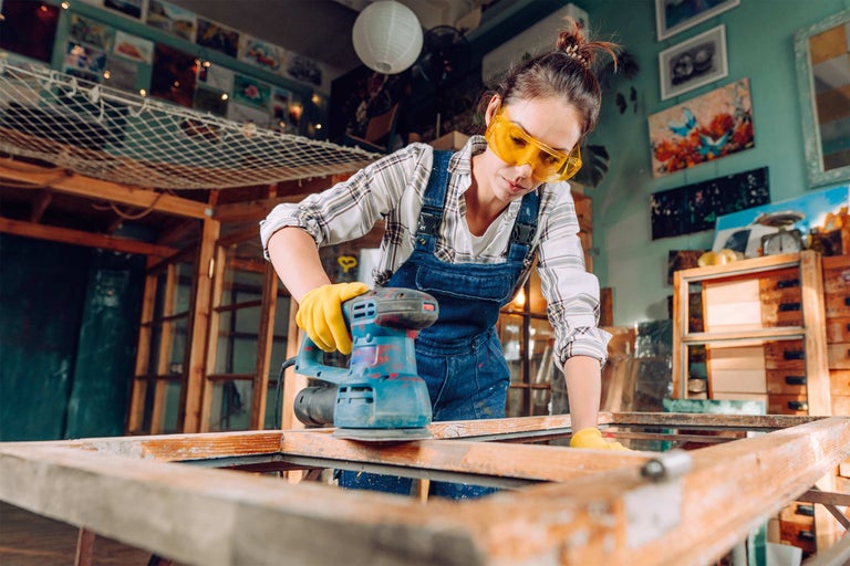 Frau schleift Holzrahmen mit einem Exzenterschleifer in einer Werkstatt. Sie trägt eine Schutzbrille und Handschuhe.