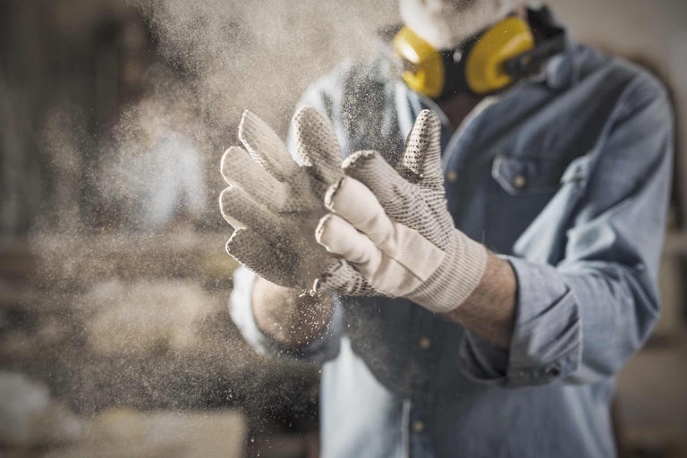 Eine Person mit Schutzhandschuhen und Ohrenschützern schlägt ihre Hände zusammen, wodurch Staub aufgewirbelt wird.