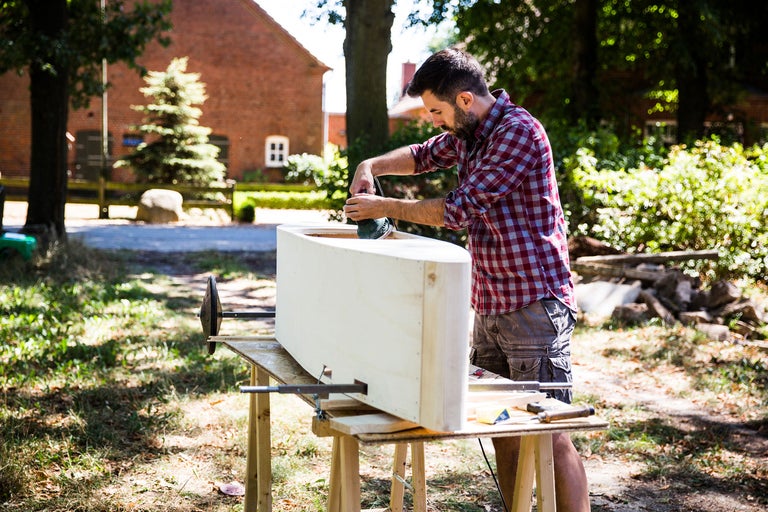 Homme ponçant une construction en bois à l'extérieur avec une ponceuse sur un établi
