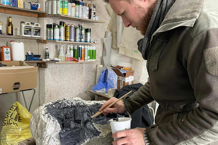 Mann bemalt Gestein mit Pinsel und Farbe in Werkstatt, Regale mit Farbe im Hintergrund.