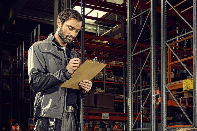 Un homme en vêtements de travail prend des notes sur un presse-papiers devant des rayonnages d'entrepôt.