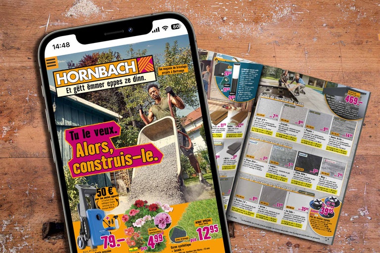 Smartphone mit Hornbach Logo und Gartenprojekten neben einem gedruckten Katalog mit Terrassenplatten und Außenleuchten auf einem Holztisch.