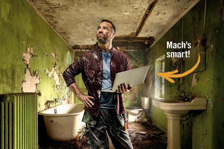 Ein Mann mit Laptop steht in einem renovierungsbedürftigen Badezimmer mit Badewanne, Waschbecken und Toilette