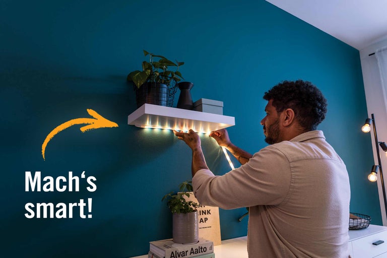 Mann befestigt LED-Streifen unter einem Wandregal mit Pflanzen und Dekorationen.