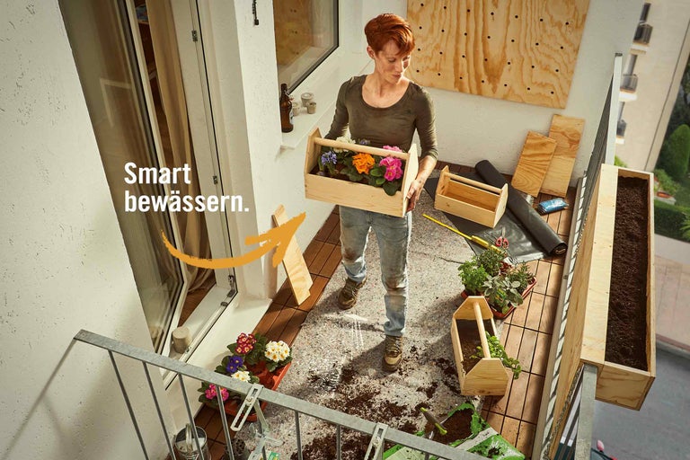 Frau bepflanzt Balkon mit Blumen und Kräutern in Holzkästen für ein smartes Bewässerungssystem.