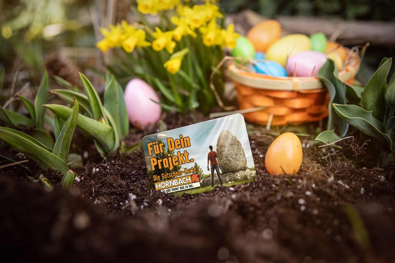 Hornbach Gutschein-Karte für Dein Projekt in einem Gartenbeet mit Erde, gelben Osterglocken und bunten Ostereiern in einem Korb.