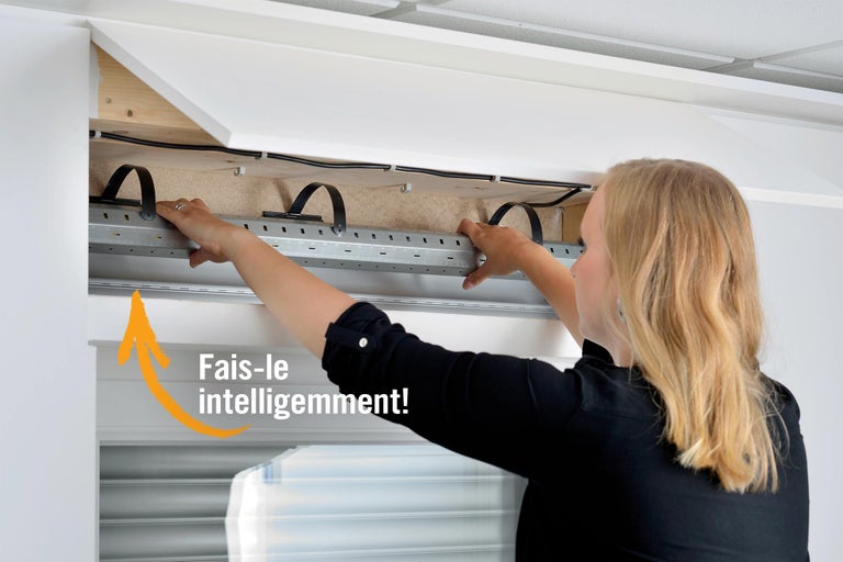 Une personne installe un axe de volet roulant dans un coffre de volet au-dessus d'une fenêtre.