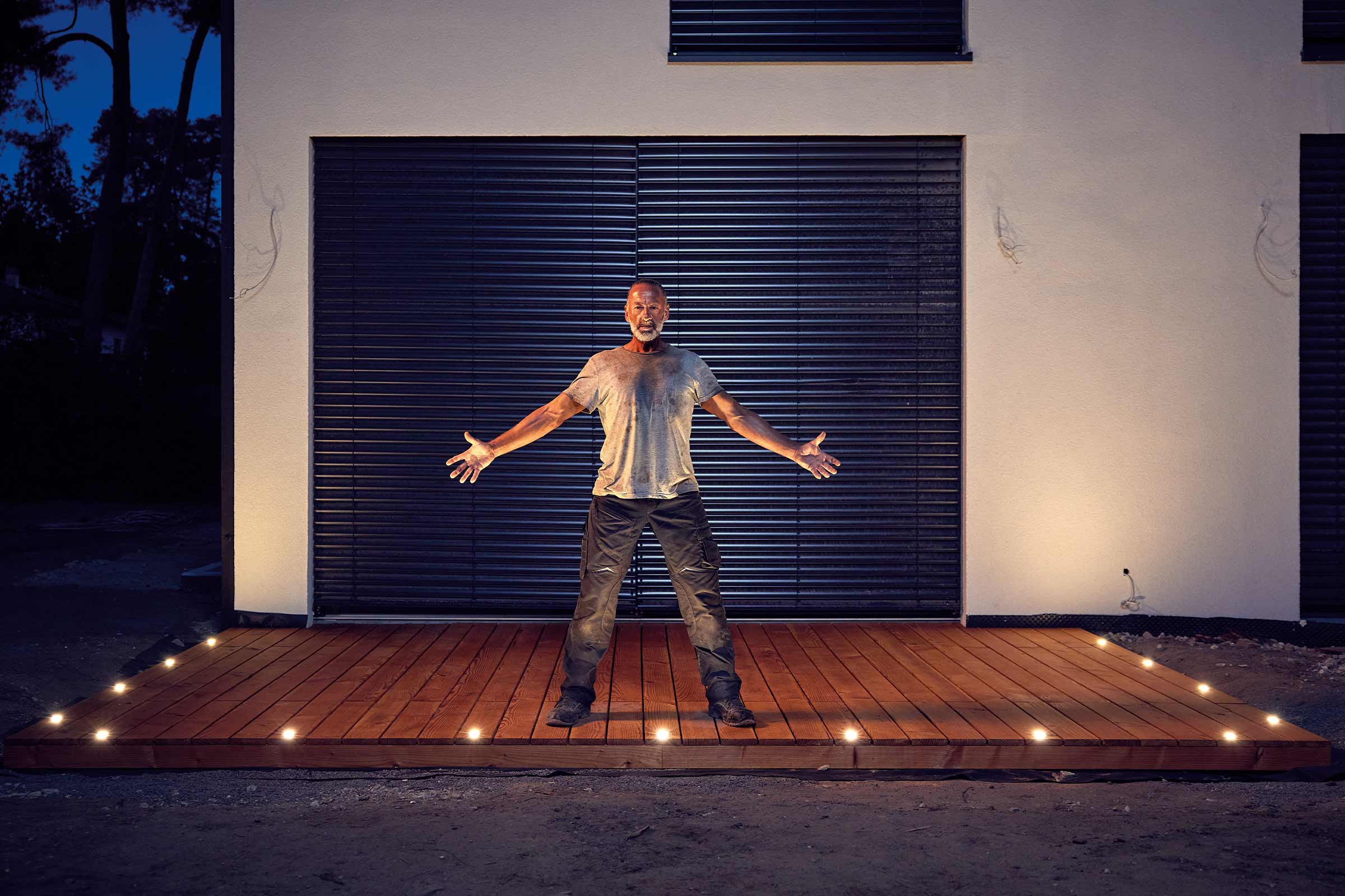 Un homme se tient sur une terrasse en bois avec des lumières intégrées devant une maison.