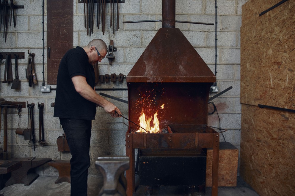Alex Pole steht in der Schmiede neben dem Feuer. Er schürt das Feuer mit einem Haken. Alex Pole steht in der Schmiede neben dem Feuer. Er schürt das Feuer mit einem Haken.