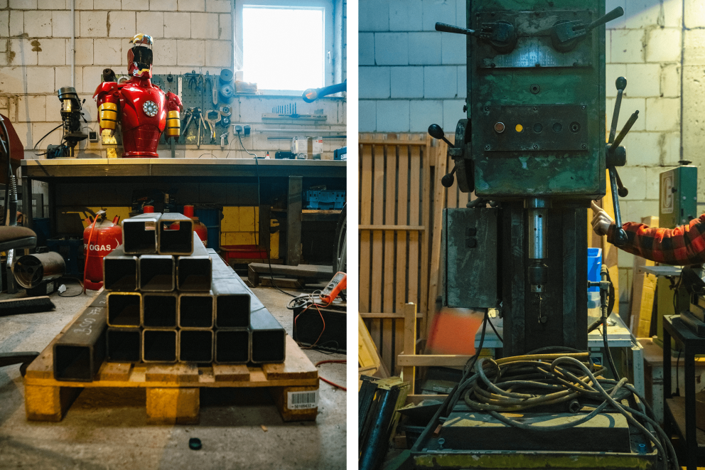 
				Palette mit Stahlträgern, dahinter Werkstatt mit „Iron Man“ Skulptur. Rechts eine alte Standbohrmaschine.

			