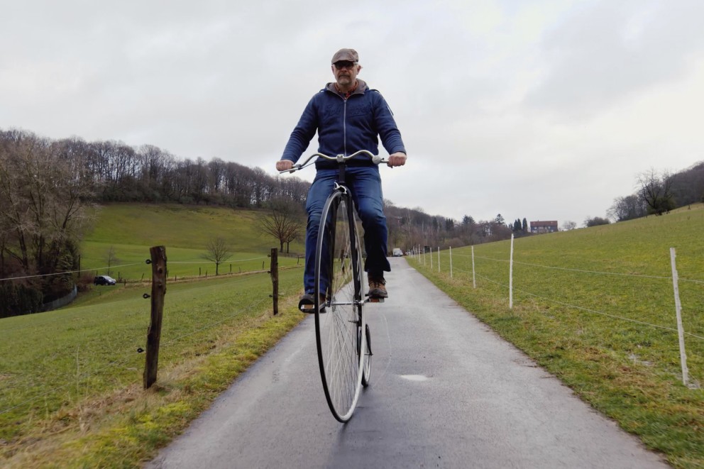 
				Bernhard fährt auf seinem Hochrad an Wiesen vorbei.

			