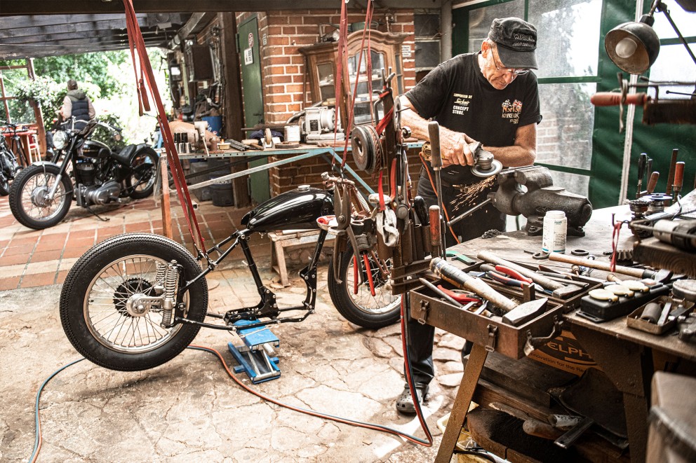 
				Mann flext ein Stück Stahl in der Werkstatt. Im Hintergrund zwei Motorräder.

			