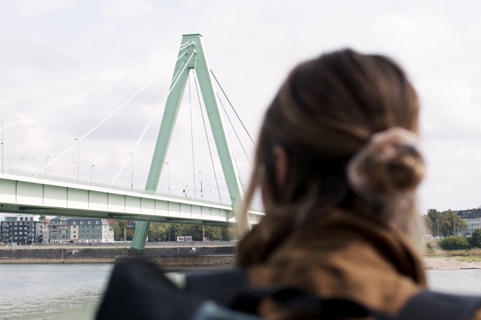 
				Anna steht am Rhein und schaut sich eine Brücke aus Stahl an

			