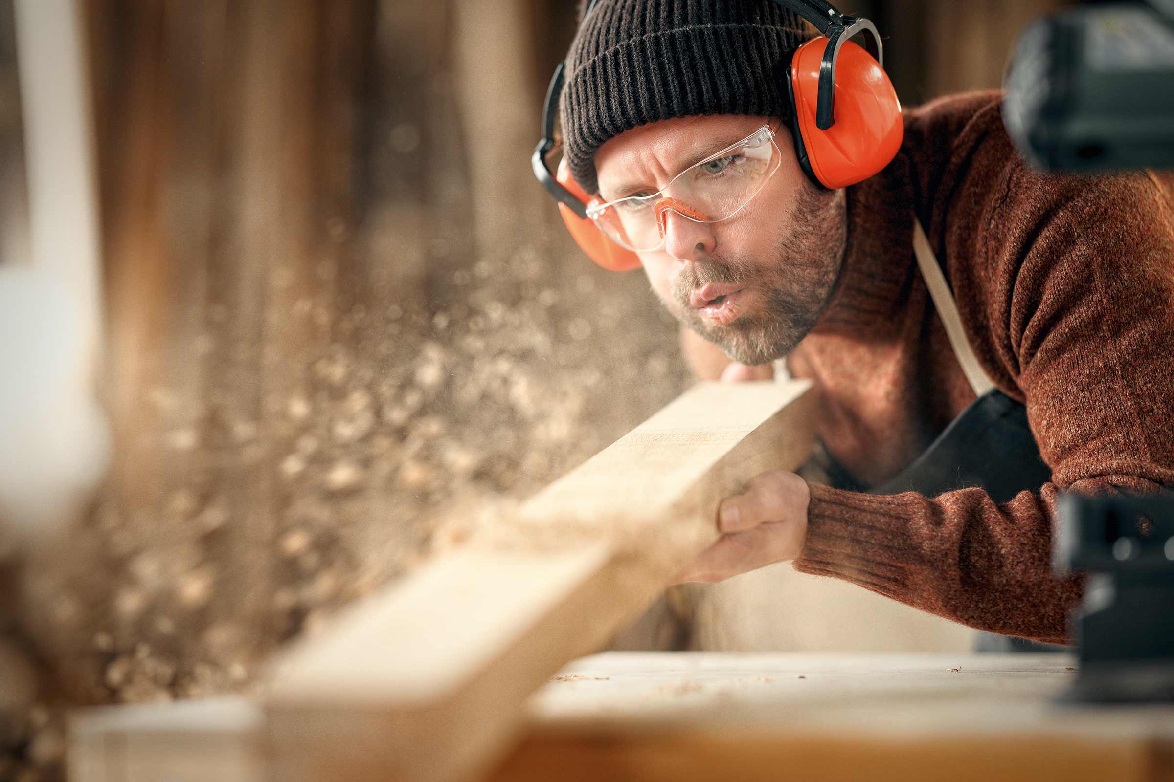 Homme soufflant de la sciure de bois d'une planche, portant des lunettes de protection et un casque antibruit