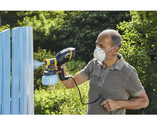 Une personne utilise un pulvérisateur de peinture pour peindre une clôture en bois. La personne porte un masque de protection.