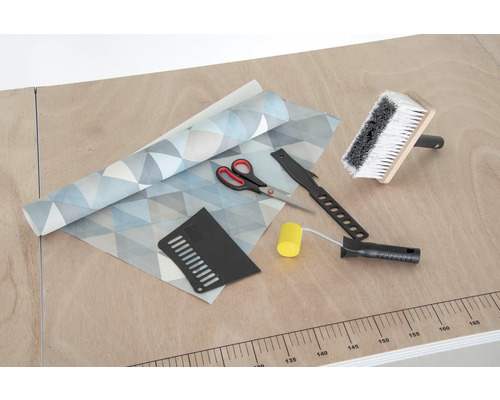 Tapete mit geometrischem Muster, Kleisterbürste, Nahtroller, Tapetenmesser, Andrückspachtel und Schere auf einem Tisch.