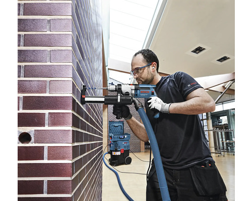 Un artisan perce un trou dans un mur de briques avec une perceuse, tandis qu''un aspirateur aspire la poussière.