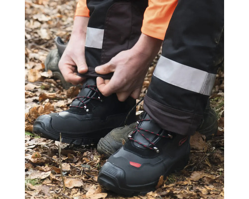 Une personne portant un pantalon de travail noir et des chaussures de sécurité se tient sur un tapis de feuilles.