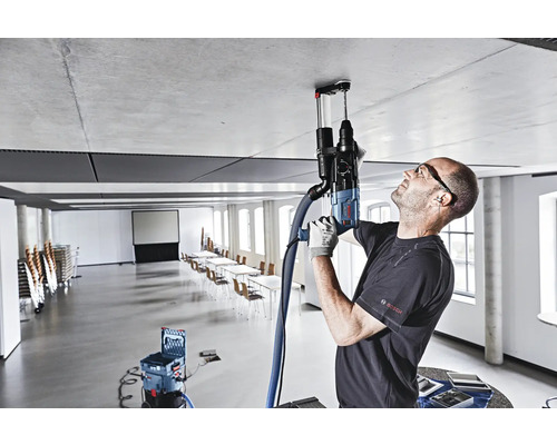 Ein Handwerker bohrt mit einem Bohrhammer ein Loch in die Decke. Ein Staubsauger ist angeschlossen, um Staub zu entfernen. Im Hintergrund befindet sich ein großer Konferenzraum.