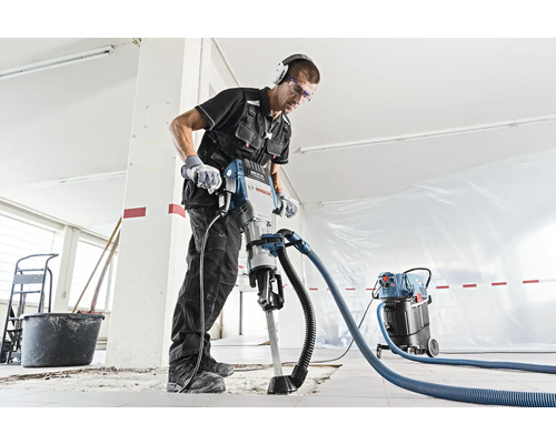 Un ouvrier du bâtiment utilise un marteau-piqueur avec extraction de poussière dans une pièce.