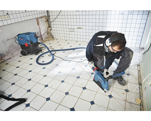 Artisan travaillant avec une rainureuse à murs et un aspirateur dans une pièce avec un sol carrelé.