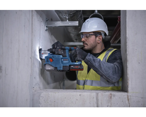 Un ouvrier du bâtiment portant un casque de sécurité et des lunettes de protection perce un trou dans un mur en béton à l'aide d'un perforateur.