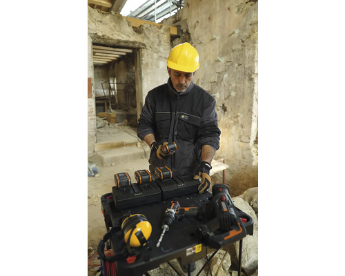 Artisan avec un casque de sécurité jaune à une table avec des batteries, des chargeurs et des outils dans un bâtiment.