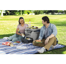 Ein Paar geniesst ein Picknick mit einem tragbaren Ninja-Grill, Speisen und Getränken auf einer blau-weiss gestreiften Decke im Freien.