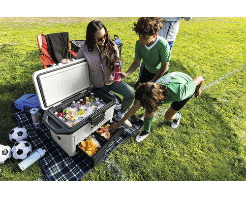 Kühlbox mit Getränken und Snacks auf einer Picknickdecke mit Fußballmotiven, umgeben von Personen auf einem Fußballfeld.