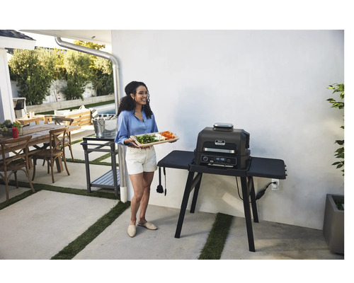 Frau mit Gemüsetablett steht neben einem Elektrogrill im Außenbereich.