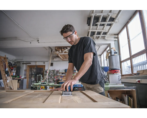 Un homme ponce une porte en bois avec une ponceuse vibrante sans fil dans un atelier.
