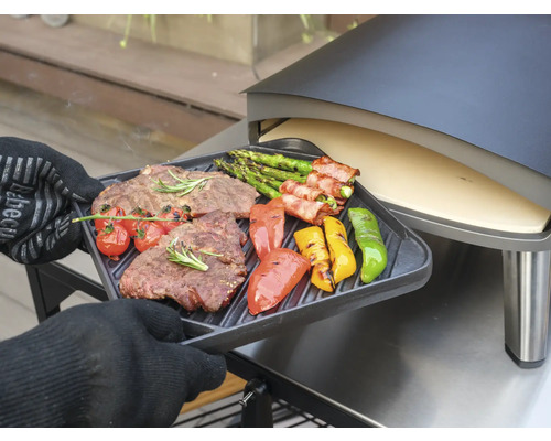 Eine Person mit Ofenhandschuhen hält ein Grilltablett mit gegrilltem Steak, Spargel, Tomaten und Paprika vor einem Pizzaofen.