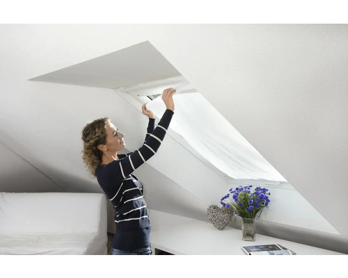 Une femme installe un store de protection solaire sur une fenêtre de toit dans une chambre mansardée lumineuse.