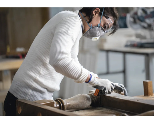 Eine Person schleift ein Möbelstück mit Schleifpapier, wobei sie eine Schutzmaske und Schutzbrille trägt.